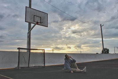 Woman sitting in sports court against cloudy sky during sunset