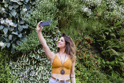 Beautiful woman taking selfie from mobile phone while standing against plants at park