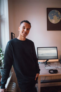 Portrait of confident young freelance worker standing against computer on desk at home