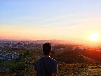 Rear view of man looking at cityscape against sky during sunset