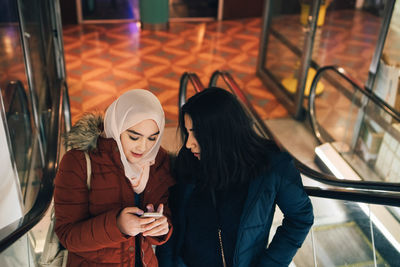 High angle view of female friends sharing smart phone while standing on escalator in shopping mall