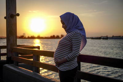 Side view of woman standing by railing against sea during sunset