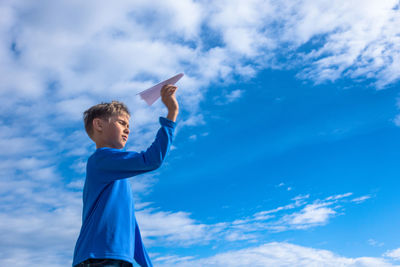 Boy holding paper airplane while standing against sky