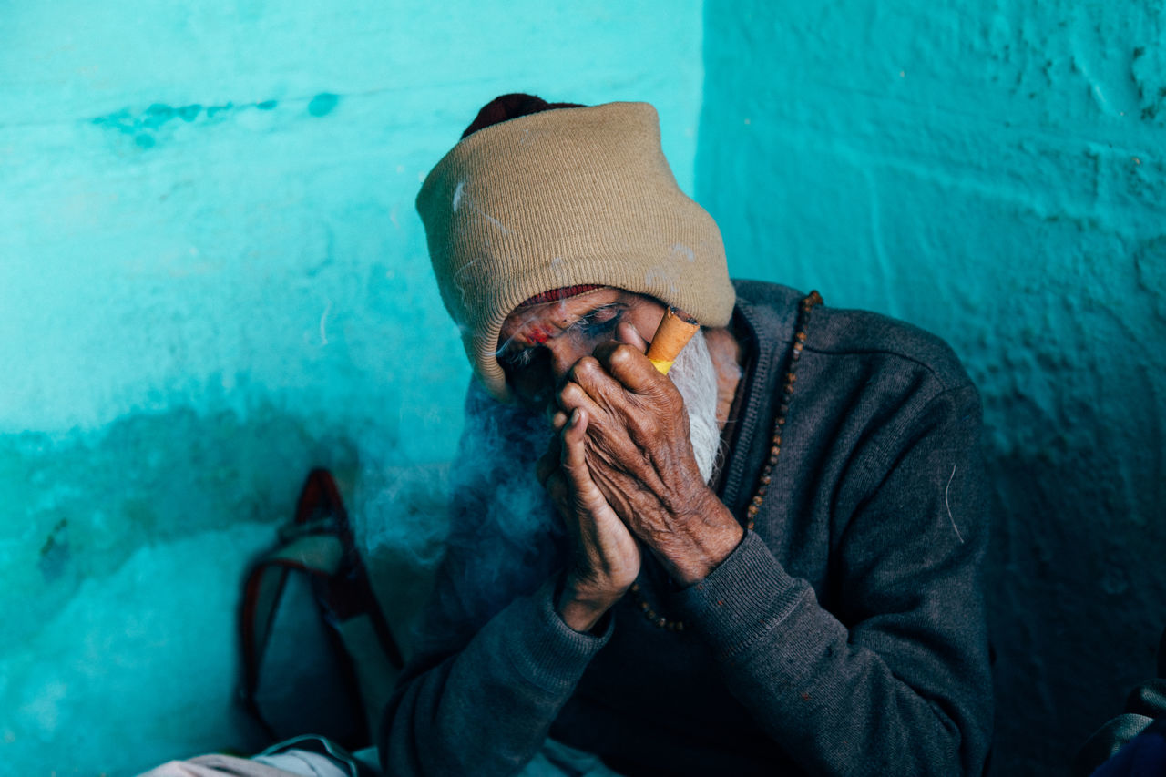 Varanasi, india - february, 2018: poor elderly hindu male in beige knitted cap sitting in corner of house with blue walls and smoking cigar
