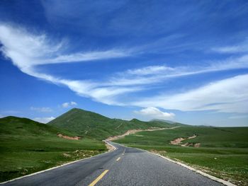 Empty road amidst green hill against cloudy blue sky