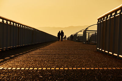 Rear view of people walking on bridge against sky during sunset