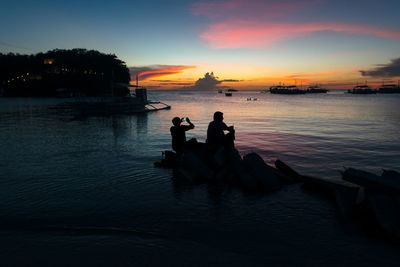 Silhouette people sitting by sea against sky during sunset