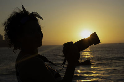 Silhouette woman with camera against sea during sunset
