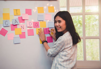 Smiling young woman standing by text over adhesive notes on whiteboard