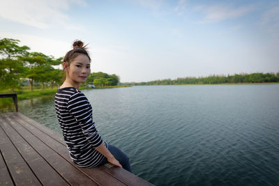 Portrait of smiling young woman sitting by lake against sky