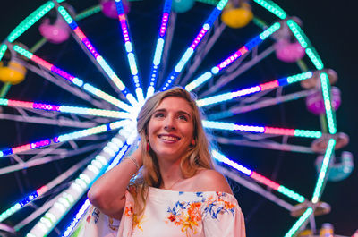 Low angle view of young woman standing against illuminated ferris wheel at amusement park