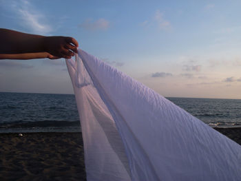 Cropped hands of person holding textile at beach against sky during sunset