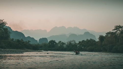 Man by river against sky during sunset