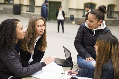 Multi-ethnic female friends discussing while studying at table in schoolyard