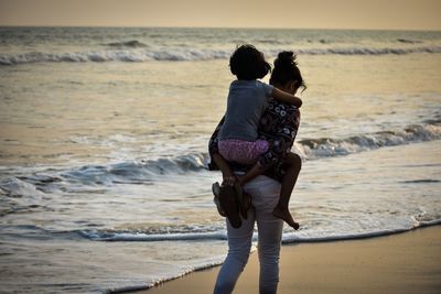 Rear view of mother giving piggyback ride to daughter at beach against sky