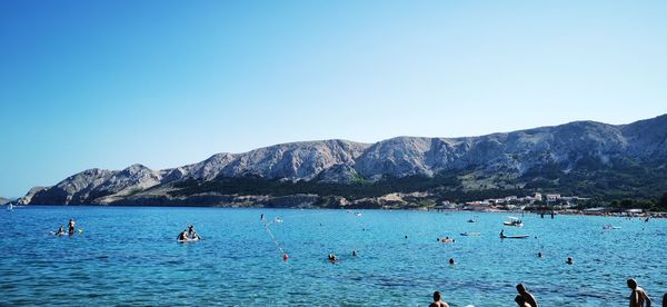 People swimming in sea by mountains against clear blue sky