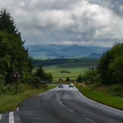 Vehicles on country road against cloudy sky