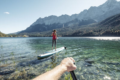 Germany, bavaria, garmisch partenkirchen, young couple stand up padling on lake eibsee, overlooking zugspitze mountain