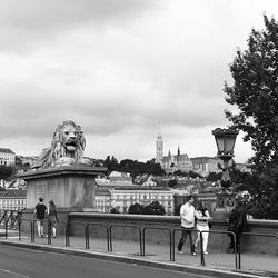 People on bridge in city against cloudy sky