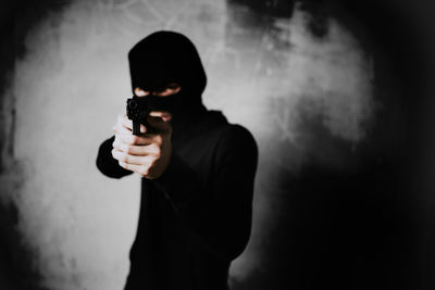 Portrait of young man aiming gun while standing against wall