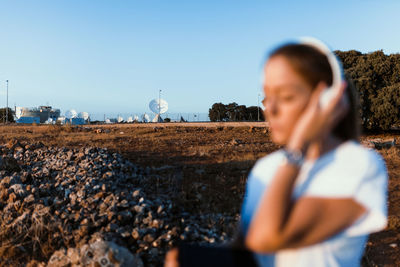 Young woman listening over headphones while sitting on land against sky