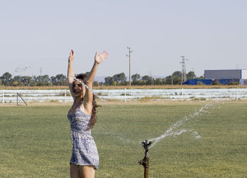 Full length of smiling young woman standing on football field, playing with sprinkler
