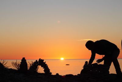 Silhouette man stacking stones at beach against orange sky during sunset