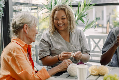 Happy young female caregiver crocheting with senior woman and man at retirement home