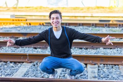 Portrait of smiling young man with arms outstretched crouching on railroad tracks