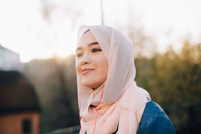 Smiling young muslim woman listening music through in-ear headphones against clear sky