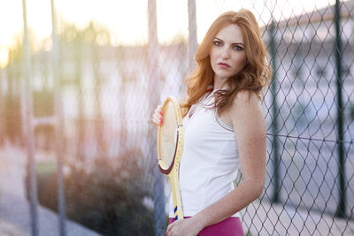 Portrait of woman holding badminton racket while standing against fence