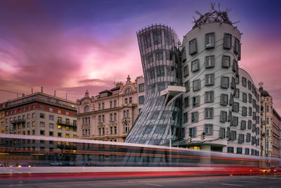 Light trails on street by buildings against sky during sunset