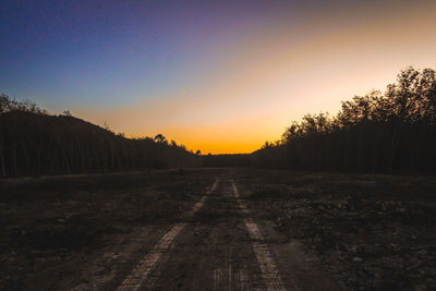 Dirt road amidst trees against clear sky during sunset