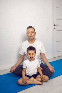 Grandmother and her grandson are sitting on a yoga mat in a white apartment at home and meditating