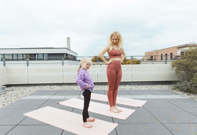 Happy mother and daughter with hands on hips practicing yoga at rooftop