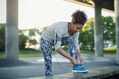 Woman tying shoelace on bench while standing at park