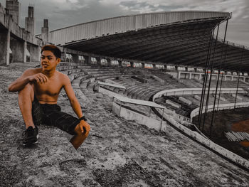 Portrait of shirtless man sitting on bridge against sky