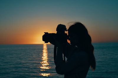 Woman photographing sea against sky during sunset