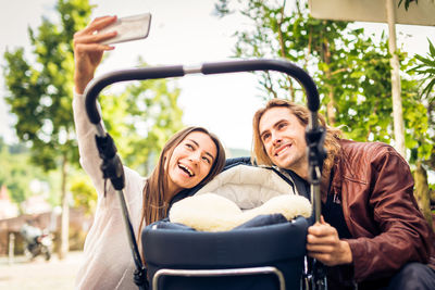 Cheerful couple doing selfie with baby outdoors