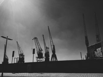 Silhouette cranes at commercial dock on sunny day against sky