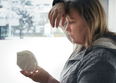 A middle aged woman stands at the window looking at an ffp 2 mask. she is worried about the future. 