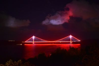 Illuminated suspension bridge over river against sky at night