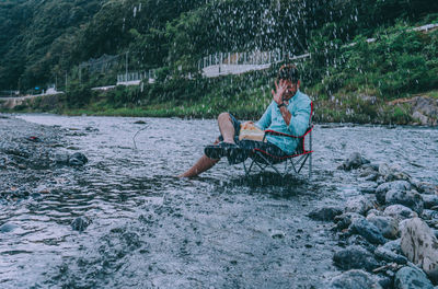 Full length of man sitting on chair in river while water splashing 