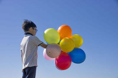 Boy holding multi colored balloons while standing against clear blue sky