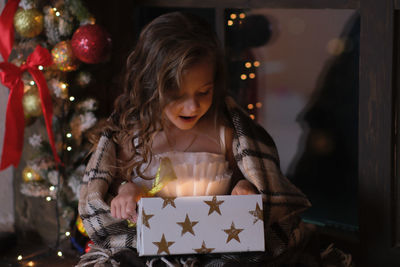 A little girl is looking at gift box and surprising near the window on christmas background.