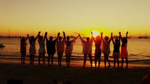 Rear view of friends holding hands with arms raised at beach during sunset