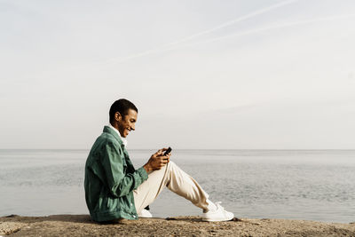 Happy man text messaging through smart phone while sitting on pier by sky and sea
