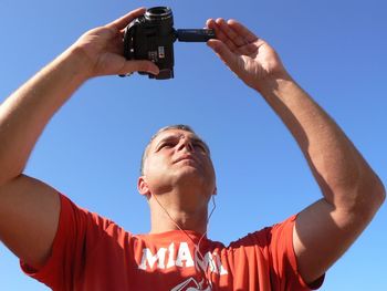 Low angle view of man using camera against blue sky during sunny day
