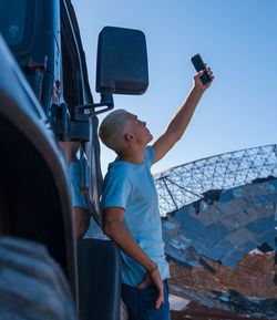 Rear view of boy photographing with mobile phone against sky