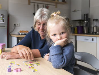 Upset blond girl sitting by grandmother playing puzzle at dining table in kitchen